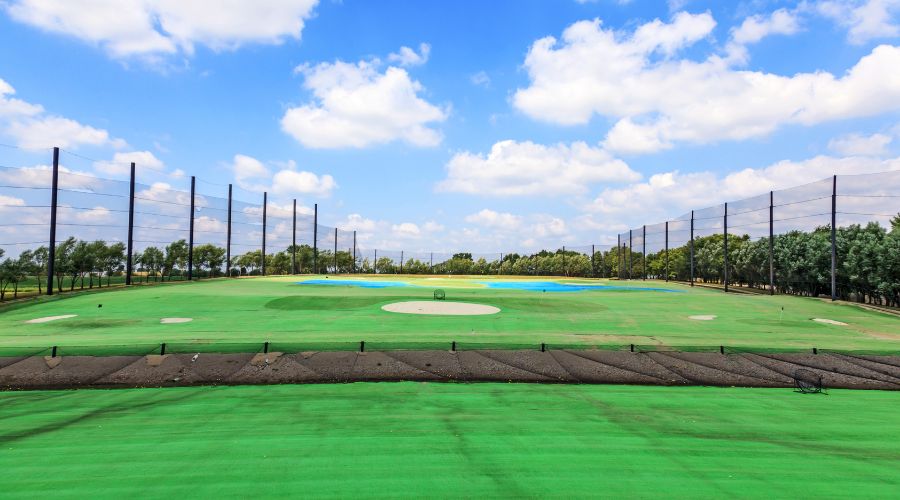 Wide view of a golf driving range with artificial turf, tall perimeter netting poles, and protective netting under a blue sky.