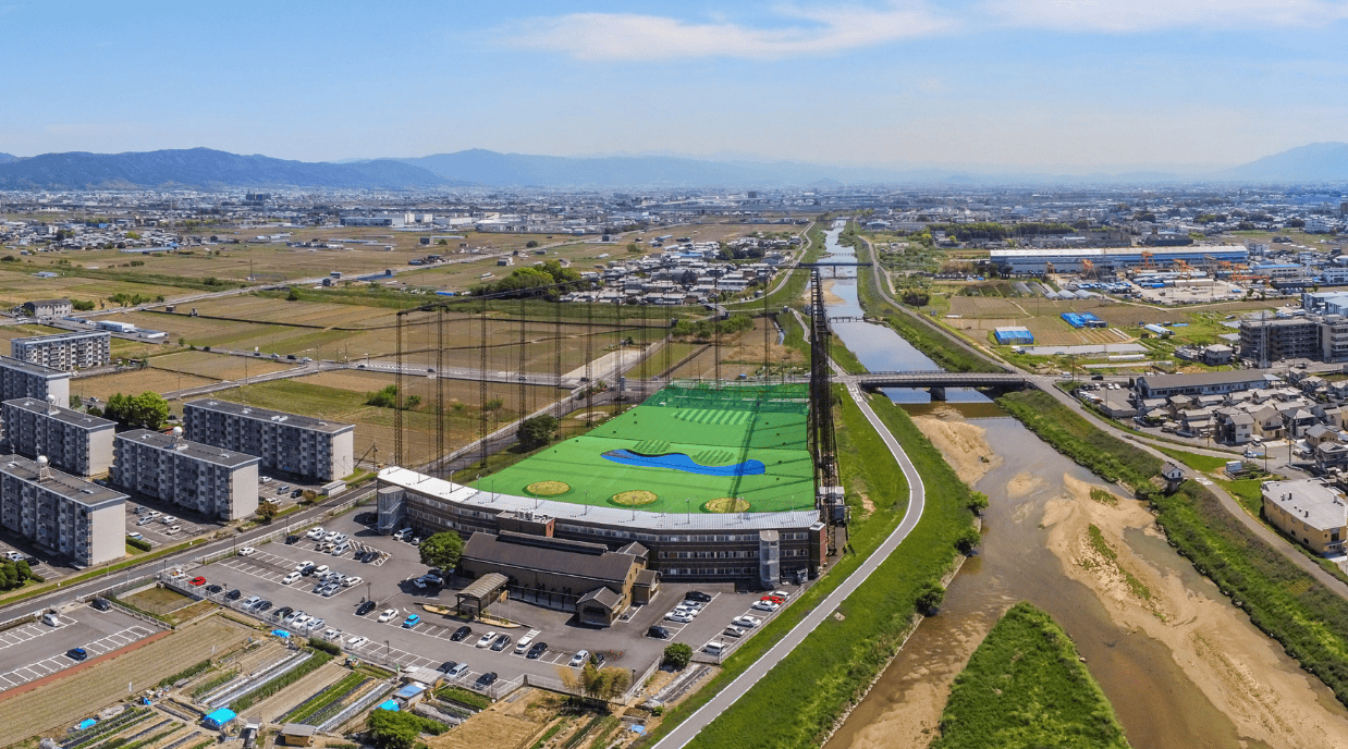 Aerial view of an urban driving range with tall perimeter netting beside apartment buildings, roads and a river.